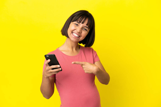 若い混合レース妊娠中の女性は、携帯電話を使用して黄色の背景に隔離し、それを指す - 写真・画像