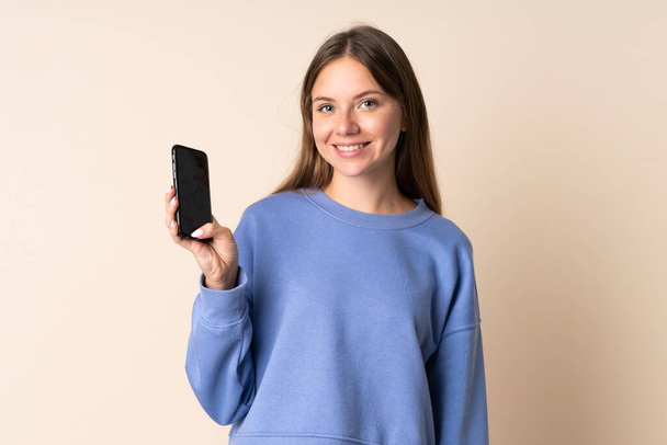Молода литовська жінка користується мобільним телефоном, ізольованим на бежевому фоні з радісним виразом. - Фото, зображення