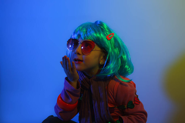 Little girl blowing kiss wearing a colorful wig and heart-shaped sunglasses posed for a photo shooting on the disco light background - Zdjęcie, obraz