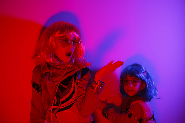 Zwei kleine Mädchen mit bunter Perücke und herzförmiger Sonnenbrille posierten für ein Fotoshooting auf dem hellen Hintergrund der Disco - Foto, Bild