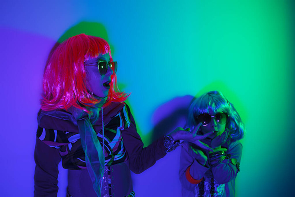 Two little girls wearing a colorful wig and heart-shaped sunglasses posed for a photo shooting on the disco light background - Photo, Image
