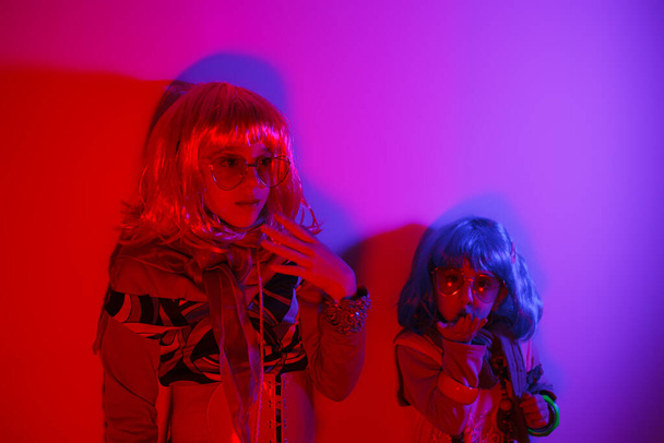 Dwie małe dziewczynki w kolorowych perukach i okularach przeciwsłonecznych w kształcie serca, pozowane do zdjęć na tle dyskotekowego światła. - Zdjęcie, obraz