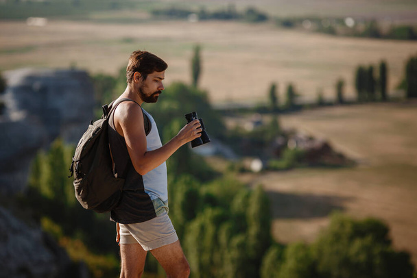 Guy looking at binoculars in hill. man in t-shirt with backpack. Young Caucasian man during hike in valley landscape - Foto, afbeelding