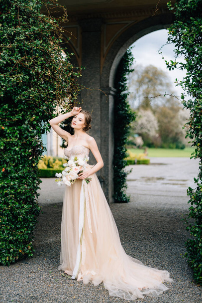 A noiva pensativa em um vestido com ombros nus mantém um buquê de flores cor-de-rosa no pano de fundo de um arco entrelaçado com vegetação. Lago de Como, Itália - Foto, Imagem