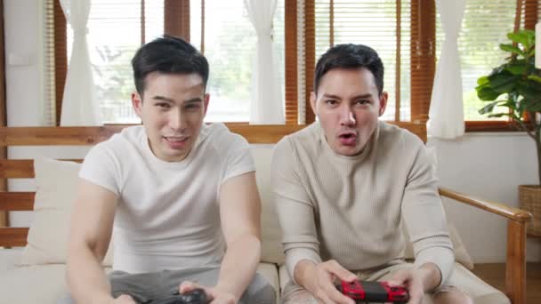 Boldog ázsiai fiatal meleg pár ül kanapén használ joystick vezérlő játék videó játék töltenek szórakoztató időt együtt a nappaliban a házban. Házas pár családi életmód, LMBTQ Pár marad otthon koncepció. - Felvétel, videó
