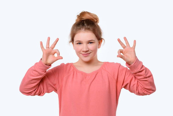 Portrait of a cute young girl in pink casual sweatshirt showing OK gesture with both hand. White background. Human emotions, gesture concept - Photo, Image