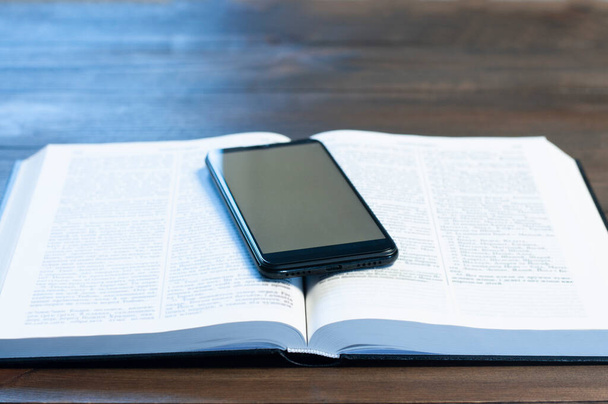 Nyitott bibliai könyv egy fa asztalon. Imádkozom. A telefon a könyvben. Üres képernyő. Munkahely az irodában. - Fotó, kép