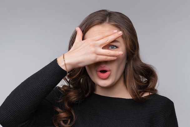 Portrait of scared and anxious young brunette woman, covering eyes with hand, peeking through fingers worried, panicking, standing on grey background - Photo, Image