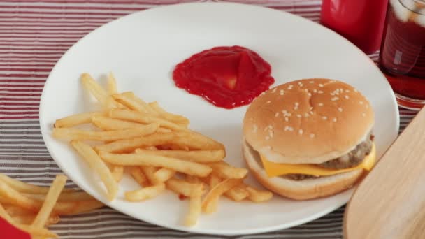 Gros plan, mise au point sélective : La main de l'homme mangeait frite, trempant du ketchup sur une assiette blanche sur une nappe rouge avec des hamburgers et du cola. Manger de la malbouffe ou de la restauration rapide pour le déjeuner est malsain. - Séquence, vidéo