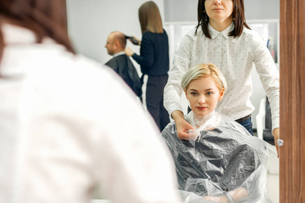 Женщина-парикмахер надевает прозрачный целлофановый плащ для клиентки в салоне красоты - Фото, изображение