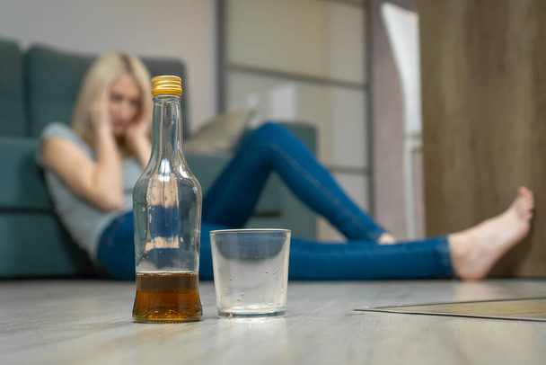 Mladá blondýnka sedí na podlaze u pohovky a drží si hlavu, v popředí je poloprázdná láhev whisky a sklenička - Fotografie, Obrázek