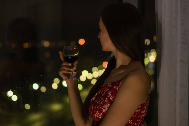 Joven mujer morena con un vestido rojo con correas y una copa de vino tinto se encuentra cerca de la ventana y mira la vida nocturna de la ciudad. Enfoque selectivo - Foto, Imagen