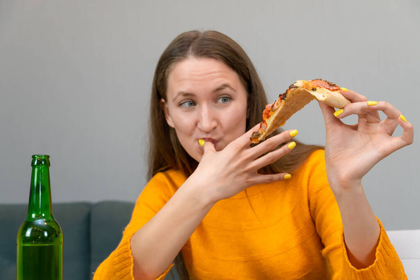 Beautiful young dark-haired woman in an orange sweatshirt is holding a piece of pizza in one hand, and the finger of the other hand is licking, next to a beer in a green bottle. Fast food concept - Photo, Image
