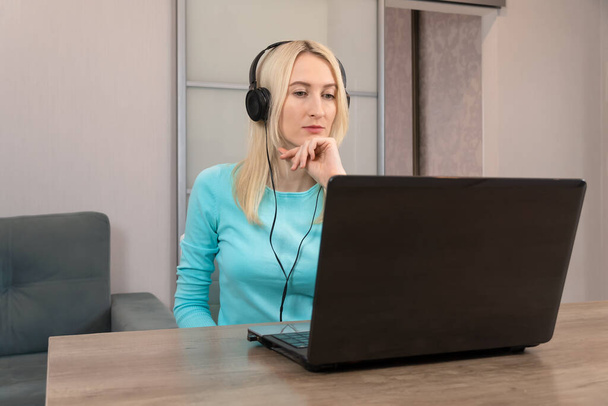 Fiatal szőke nő fülhallgatóval ül az asztalnál, laptopot néz és figyelmesen hallgat. - Fotó, kép