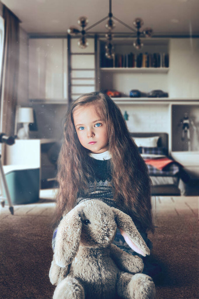Девочка сидит в комнате, а перед ней игрушечный заяц. Девушка с длинными вьющимися волосами, голубыми глазами. Плюшевая игрушка. кролик - Фото, изображение