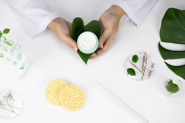 Handen houden crème pot op witte laboratoriumachtergrond, plantaardige cosmetica - Foto, afbeelding