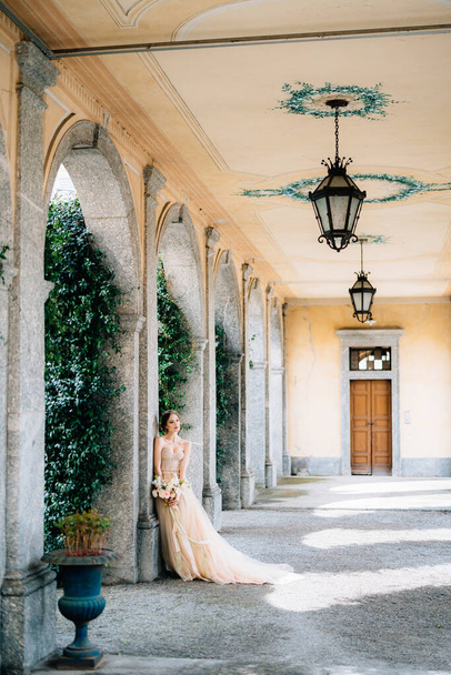 Beautiful bride in a dress with a bouquet of pink flowers is leaning against a pillar in the vaulted room. Lake Como, Italy - Photo, Image