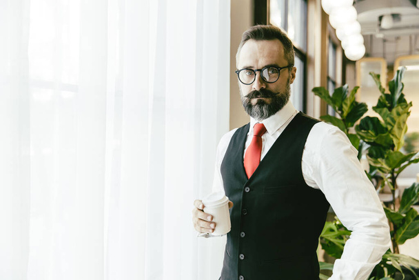 Portrait serious professional conservative adult model businessman Latin beard standing hand holding coffee cup in modern office with copy space for text. - 写真・画像