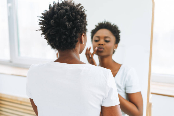 Portret van gelukkige mooie jonge Afro-Amerikaanse vrouw in wit t-shirt neemt make-up tegen spiegel - Foto, afbeelding
