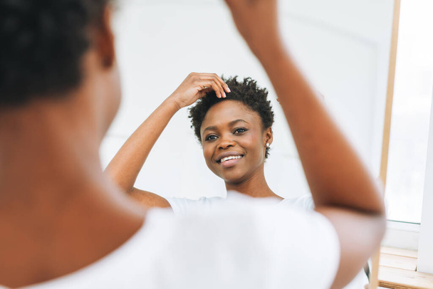 Porträt der glücklichen schönen jungen Afroamerikanerin im weißen T-Shirt macht Frisur in Spiegelnähe - Foto, Bild