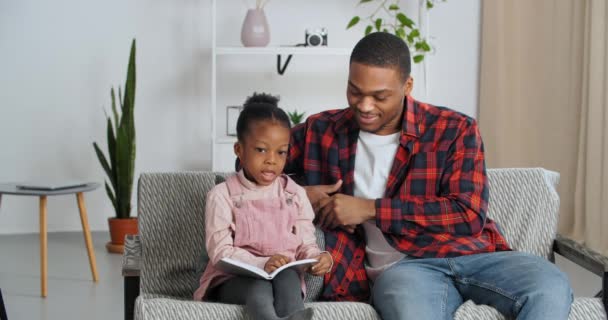 Afro-americano padre single e bambina prescolare seduti insieme sul divano in salotto a fare i compiti, il giovane padre amorevole aiuta la sua adorata figlia con lezioni insegna al bambino a leggere il libro - Filmati, video