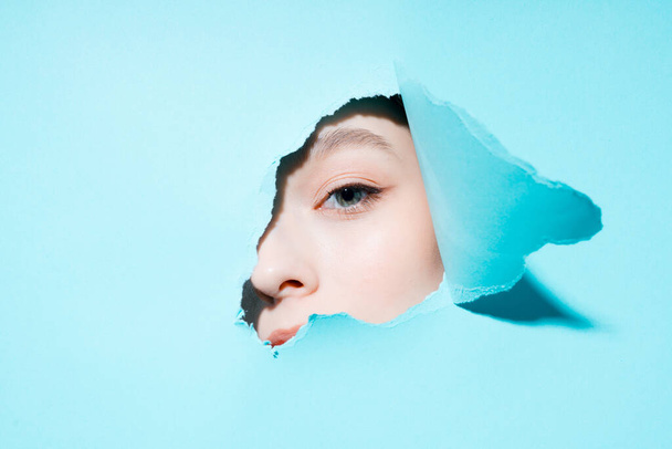 La mitad femenina de la cara mira a la cámara a través de un agujero en un fondo azul - Foto, Imagen