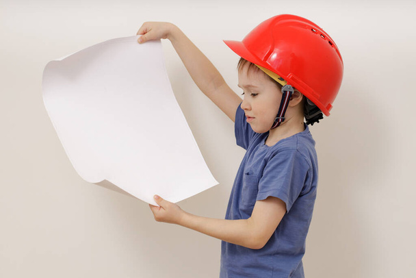 cute, charming Caucasian boy in a five-year-old hard hat holds a large project sheet in his hands. - Fotó, kép