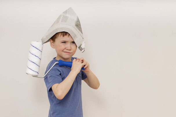 Очаровательный маленький эмоциональный 5-летний мальчик в шляпе газеты держит в руках белый роллер. концепция жилого дома (ремонт). копировальное пространство - Фото, изображение