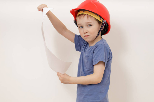 Leuke charmante blanke jongen in een helm van vijf jaar oud houdt een groot projectblad in zijn handen. - Foto, afbeelding