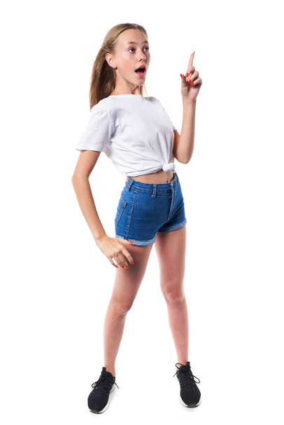 Full length of surprised teen girl wearing white t-shirt and denim shorts pointing up at blank copy space, over white background - Photo, image
