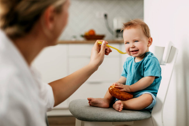 Mom feeds her little son sitting on a chair in the kitchen - Photo, Image