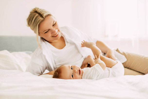 Happy mom plays with her little son lying on the bed, mom and baby relax together. Innocence, unity and family - Foto, imagen