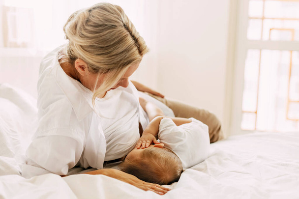 Egy fiatal anya szoptatja újszülött gyermekét, miközben az ágyon fekszik. Gondozás, gyermekkor, anyaság - Fotó, kép