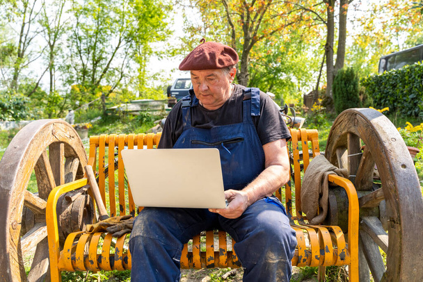 portrait of mature blacksmith. Worker on the computer outdoor sitting on a bench in the countryside  - Фото, изображение