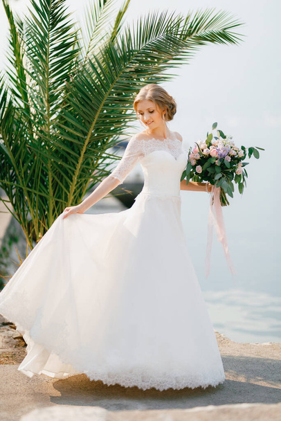 Eine zarte Braut steht in der Nähe einer Palme und hält einen Brautstrauß und den Rand ihres Rockes  - Foto, Bild