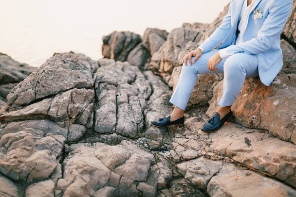 Мужчина в синем костюме с бутоньером и мокасином сидит на скалах  - Фото, изображение