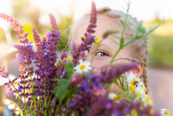 Retrato de cerca de la pequeña niña rubia caucásica linda espiando y escondiéndose sosteniendo ramo de salvia salvia flores silvestres caminando en el prado de hierba al aire libre el día de verano. Joven mujer flores silvestres - Foto, imagen