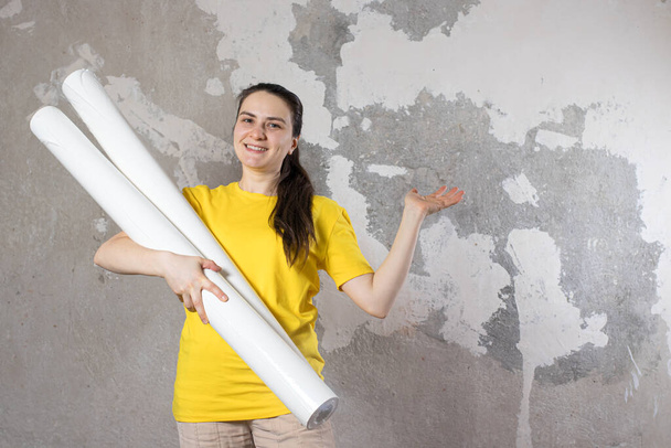 Uma mulher em uma camiseta amarela brilhante segura 2 rolos de papel de parede branco contra a parede velha. Reparação, papel de parede. Com um lugar para texto para copiar espaço. - Foto, Imagem