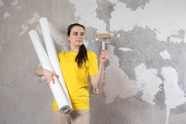 Parlak sarı tişörtlü bir kadın iki rulo beyaz duvar kağıdını eski duvara dayadı. Tamir işleri, duvar kağıtları. Metin kopyalamak için bir yer ile. - Fotoğraf, Görsel