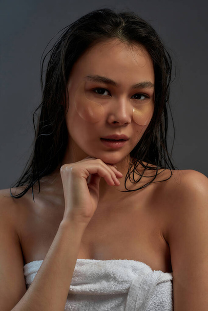 Portrait of a young asian half naked woman in bath towel with cosmetic eye patches on face looking at camera while posing against dark background - Фото, зображення