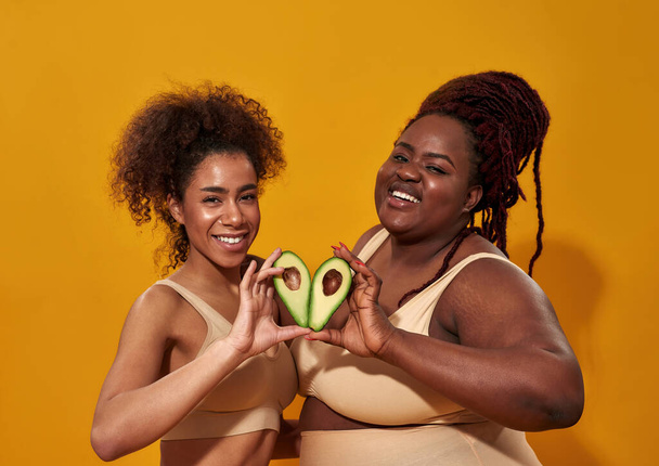 Dos mujeres afroamericanas en ropa interior sonriendo ante la cámara, sosteniendo mitades de aguacate de frutas saludables como un signo del corazón, posando aisladas sobre un fondo amarillo - Foto, imagen