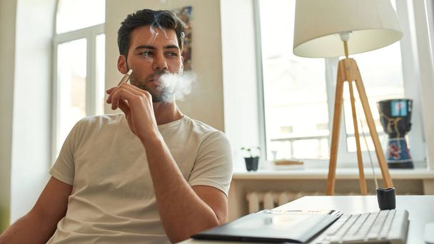 Junger Mann raucht elektronische Zigarette und schaut auf Bildschirm  - Foto, Bild