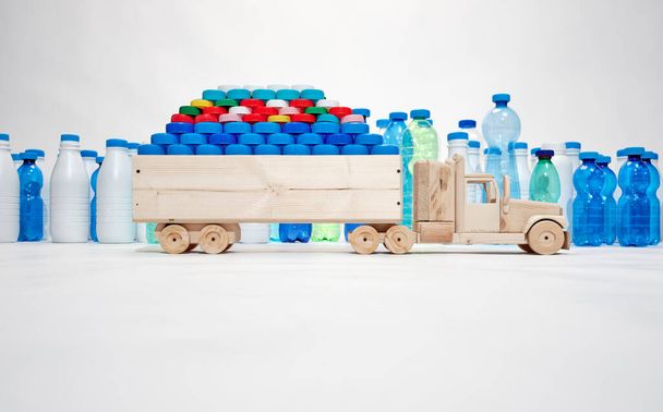 Dřevěný náklaďák nese plastové uzávěry, plastové láhve v pozadí. Plastová koncepce města. Koncept bez plastů. Koncept dopravy s nízkými emisemi CO 2. - Fotografie, Obrázek