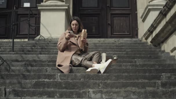Młoda stylowa kobieta jeść fast food na zewnątrz - Materiał filmowy, wideo