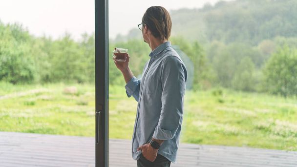 Side view of young businessman in glasses holding cup of coffee and standing near the window - Photo, Image
