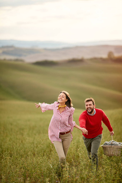 A young cheerful couple running on a large meadow while holding by hands on a beautiful day. Love, relationship, together, nature - Fotografie, Obrázek
