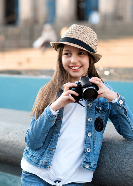 Chica vistiendo abrigo de mezclilla, sombrero de paja y jeans con cámara caminando por el centro de la ciudad y tomando fotos - Foto, Imagen