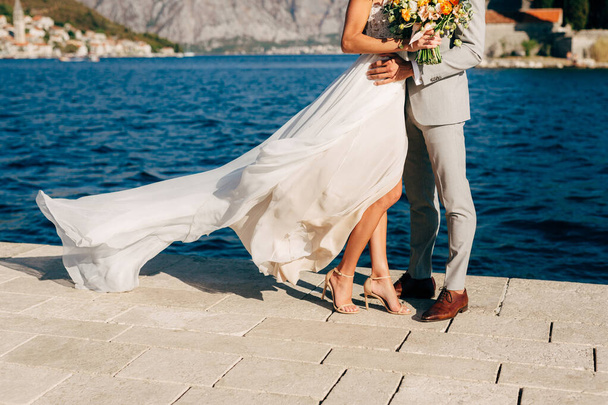 Benen van de bruid en bruidegom knuffelen op de pier in de baai van Kotor achter hen de oude stad Perast  - Foto, afbeelding