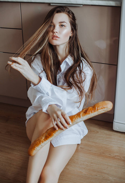 A woman in a white shirt sits on the kitchen floor next to the headset holding a long baguette in her hand. - Valokuva, kuva