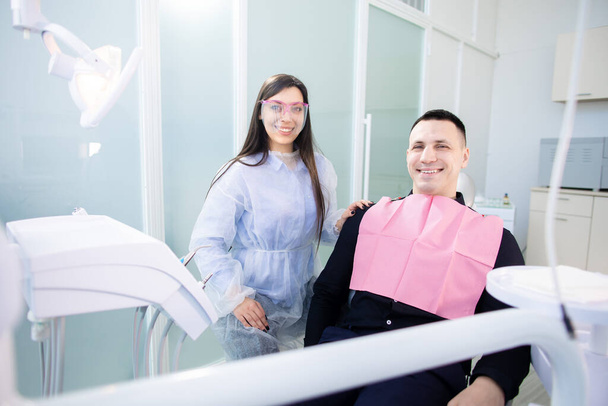 Un joven y una doctora en el consultorio de dentistas, sentados y sonriendo, mirando a la cámara. Mujer sosteniendo una herramienta de tratamiento dental - Foto, Imagen